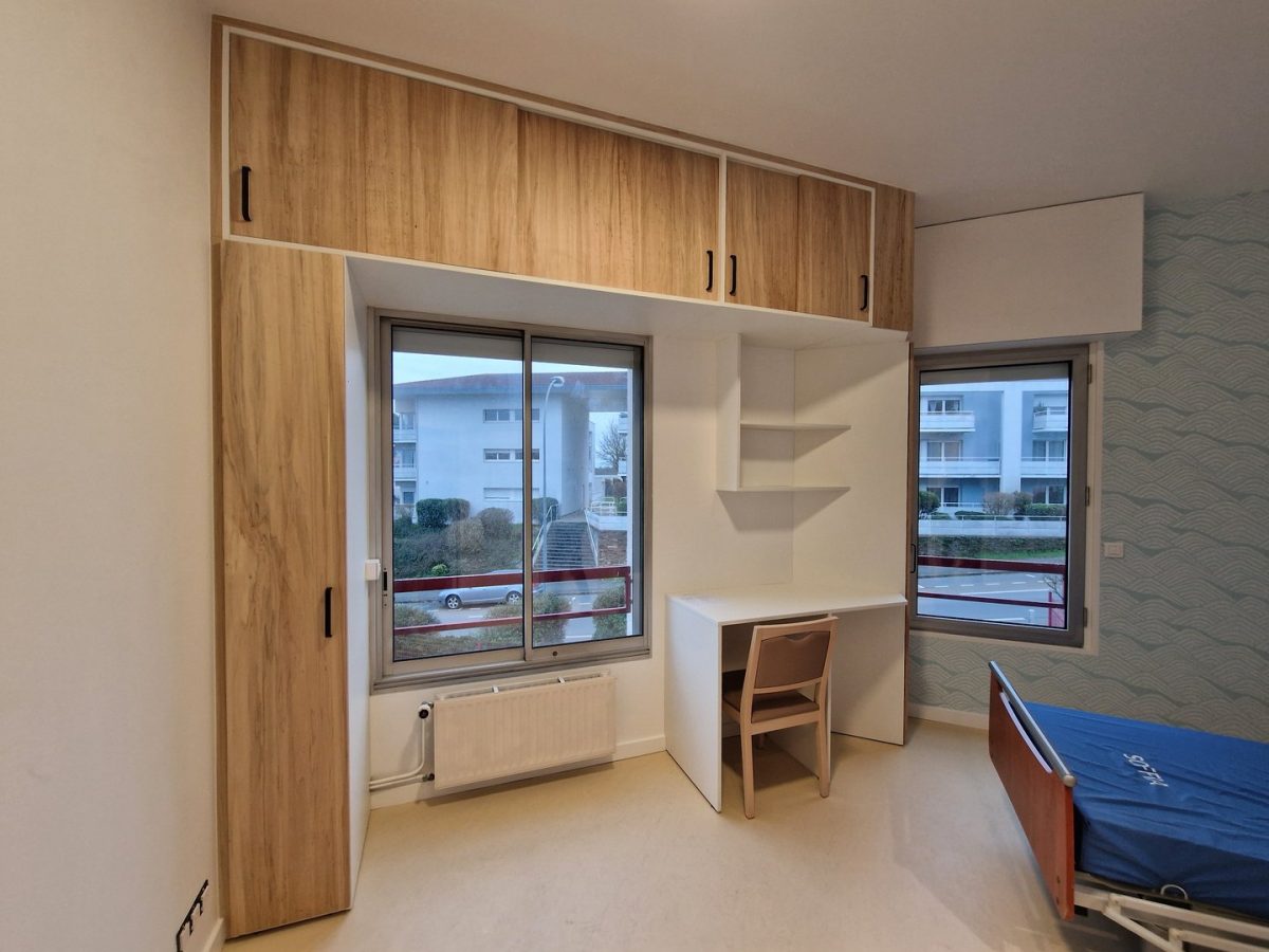 Chambre moderne avec bureau intégré, rangements bois et blanc, deux fenêtres donnant sur résidence