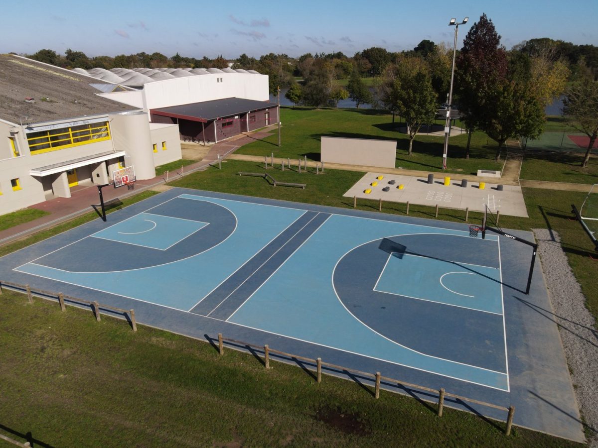 Terrain de basketball extérieur bleu avec bâtiment scolaire moderne blanc et jaune, espaces verts et équipements sportifs