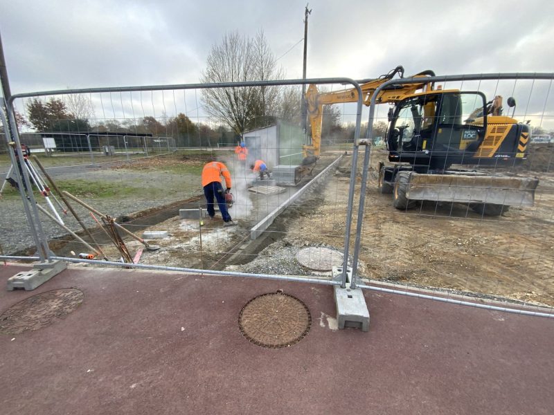 Chantier de construction avec clôture temporaire, ouvriers en gilets orange et pelleteuse jaune travaillant sur fondations