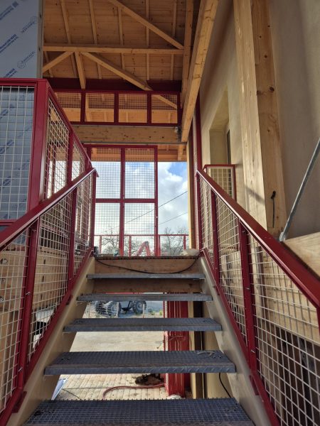 Escalier intérieur en acier rouge avec marches caillebotis, garde-corps grillagé, charpente bois apparente et mezzanine