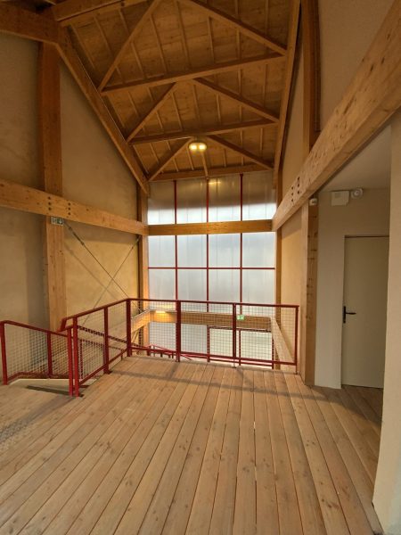 Mezzanine en bois avec garde-corps métallique rouge, charpente apparente et grande baie vitrée à cadre rouge