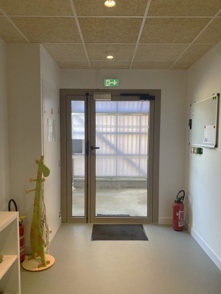 Entrée moderne avec porte vitrée encadrée d'aluminium, plafond acoustique et signalétique de sortie de secours