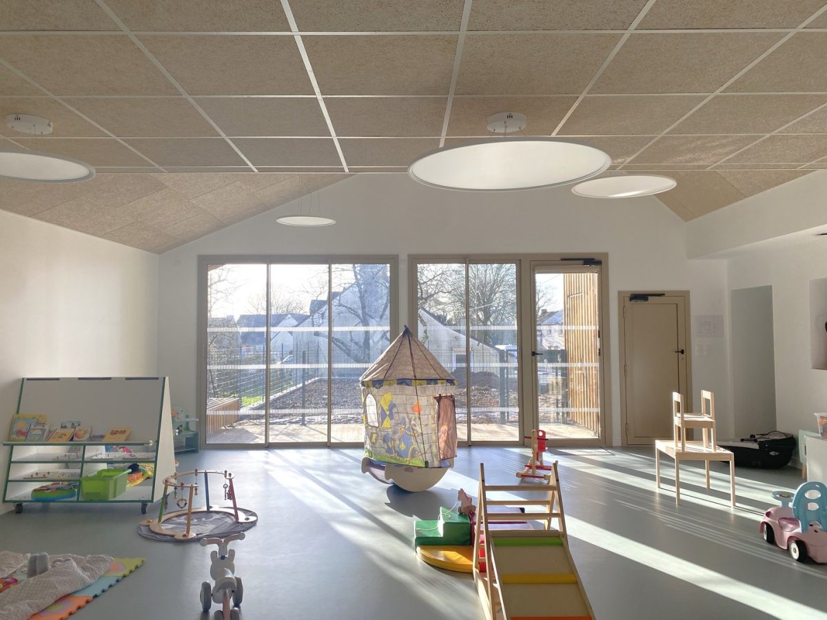 Salle de jeu lumineuse pour enfants avec plafond acoustique, luminaires circulaires et grandes baies vitrées donnant sur l'extérieur