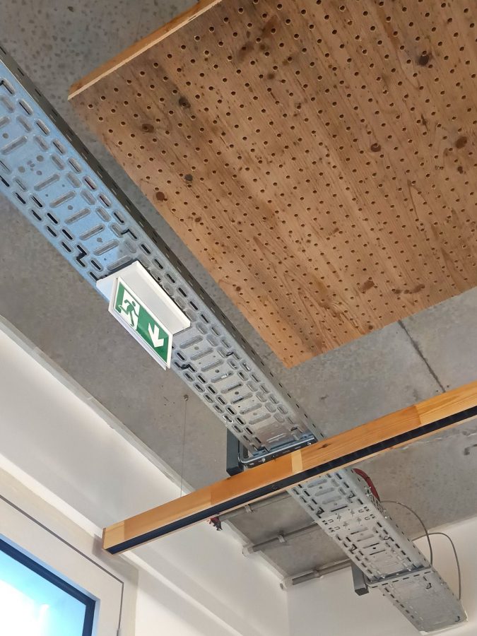 Plafond en béton brut avec panneau acoustique bois perforé, conduits techniques apparents et signalétique issue de secours