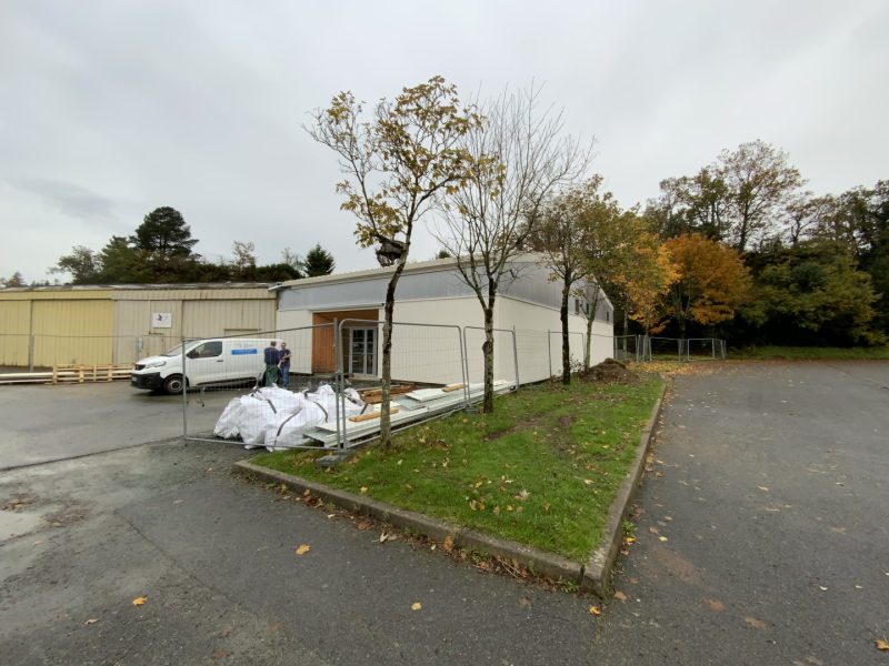 Bâtiment industriel à bardage métallique blanc avec entrée vitrée, en cours de rénovation, entouré d'arbres d'automne