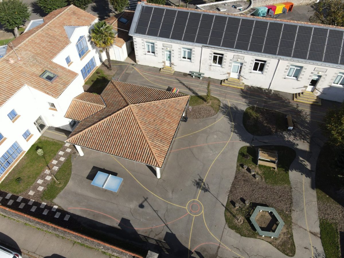 Vue aérienne d'une école avec bâtiments à toits de tuiles, panneaux solaires, cour aménagée avec jeux et marquages au sol