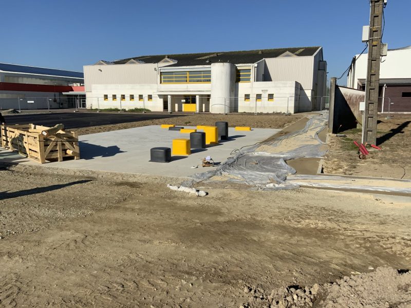 Chantier en cours avec dalle bétonnée, barrières jaunes et noires, bâtiment industriel blanc en arrière-plan