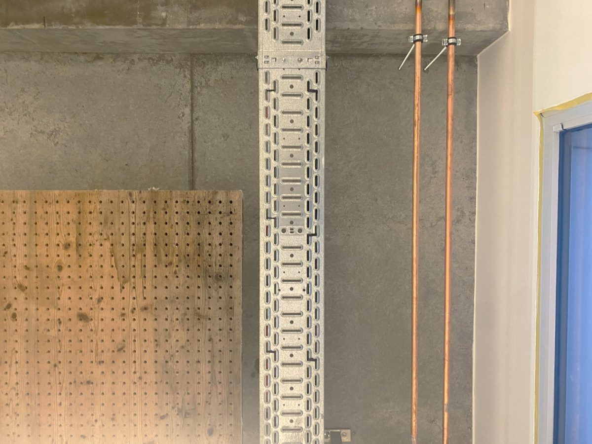 Détail de mur en béton brut avec panneau perforé en bois, rail métallique vertical et tuyaux en cuivre apparents