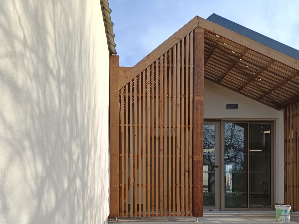 Bâtiment moderne avec façade en claire-voie bois verticale, pergola et baies vitrées, mur blanc adjacent