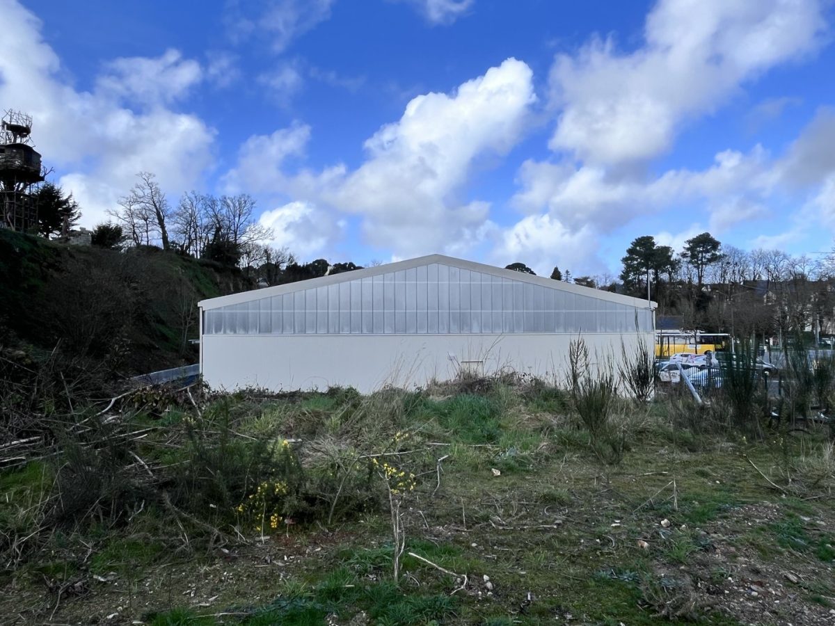 Serre moderne à structure métallique et panneaux translucides, implantée sur un terrain en friche avec végétation sauvage