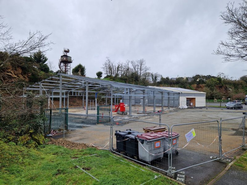 Chantier de construction avec structure métallique apparente, bardage blanc en cours d'installation, site en pente arboré