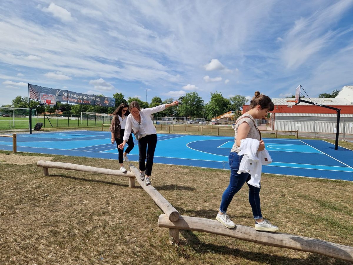 Terrain multisport bleu avec équipement d'équilibre en bois au premier plan, bâtiments et espaces verts en arrière-plan