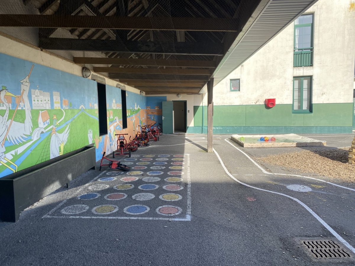 Préau d'école avec mural coloré, jeux au sol et tricycles, structure couverte donnant sur cour et bâtiment vert et blanc