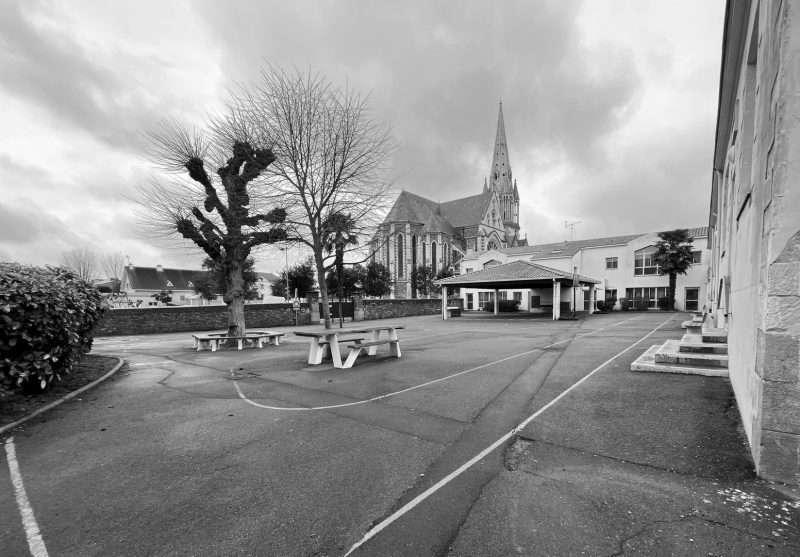 Cour d'école avec église néogothique en arrière-plan, arbres taillés et tables de pique-nique, bâtiments résidentiels