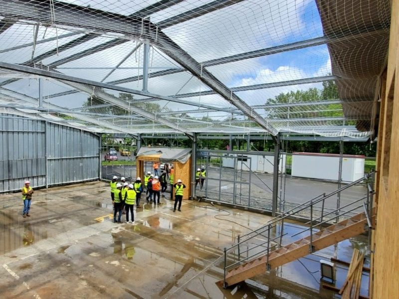 Bâtiment agricole en construction avec structure métallique, toit transparent et personnel en gilets jaunes sur dalle béton