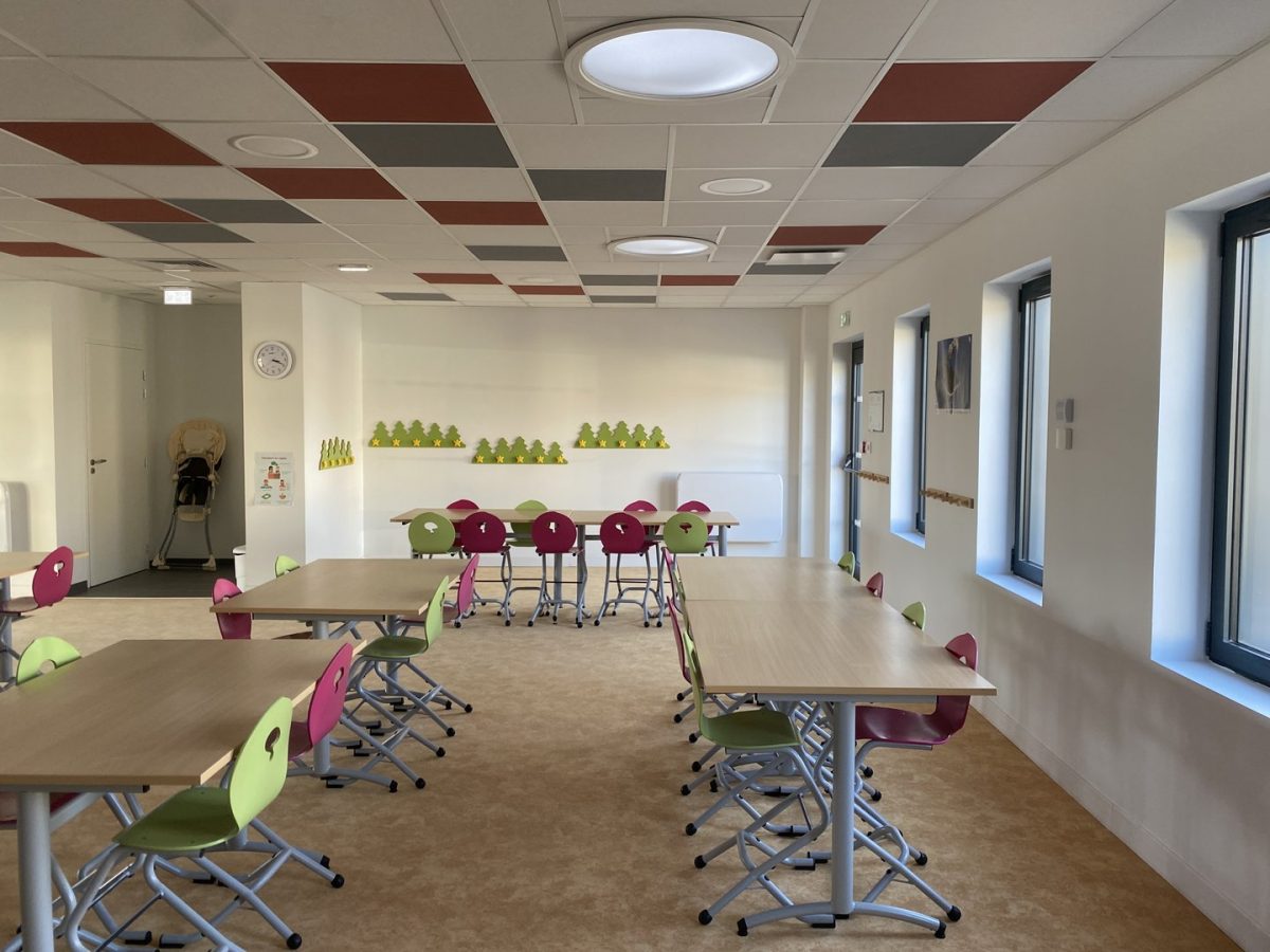 Salle de classe moderne avec tables en bois, chaises colorées, plafond acoustique décoratif et fenêtres lumineuses