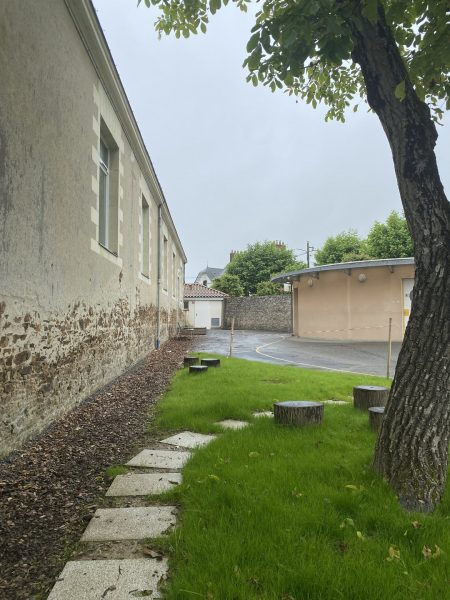 Cheminement paysager avec rondins de bois et dalles sur pelouse, entre bâtiment ancien aux murs en pierre apparente et rue