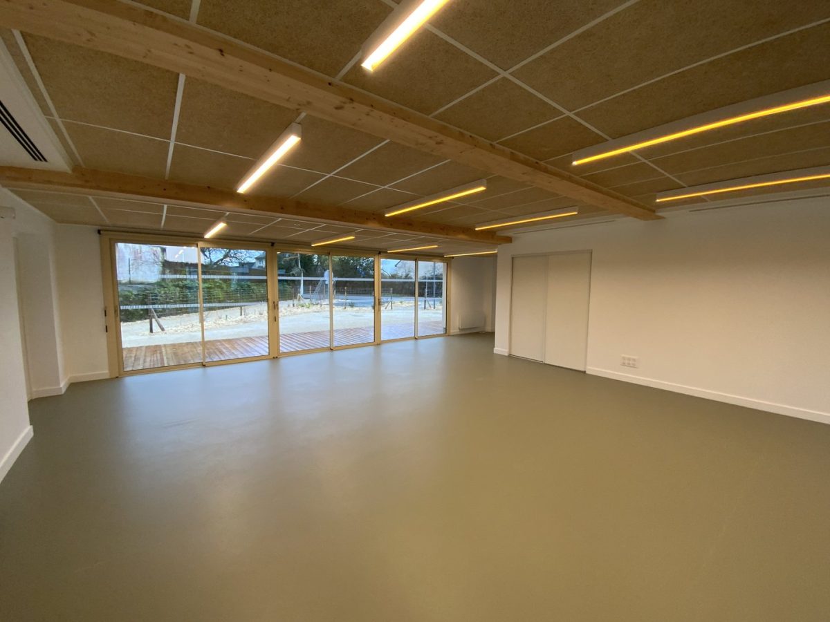 Salle vide avec plafond bois décoratif, éclairage linéaire intégré, baies vitrées donnant sur cour extérieure