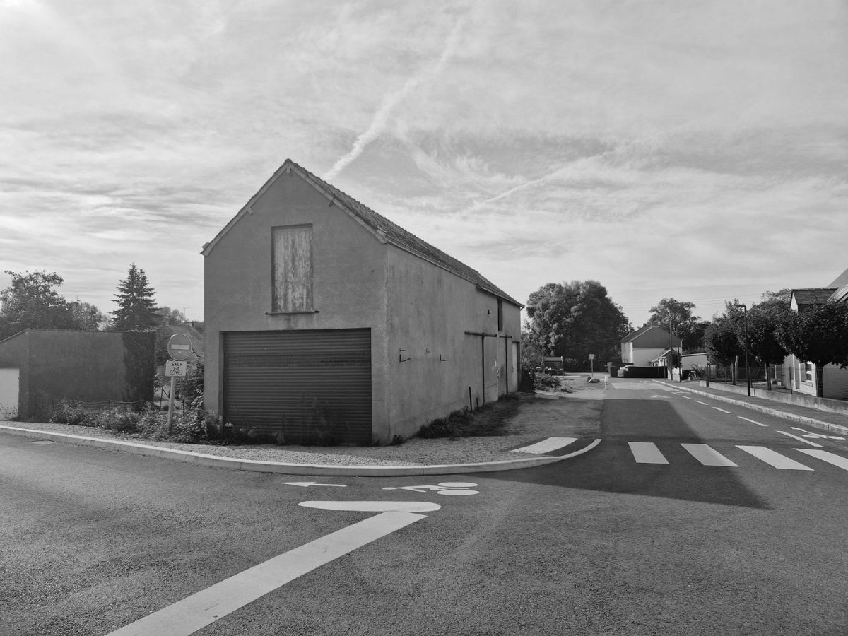 Bâtiment ancien en crépi gris avec porte de garage métallique, situé à l'angle d'une rue résidentielle