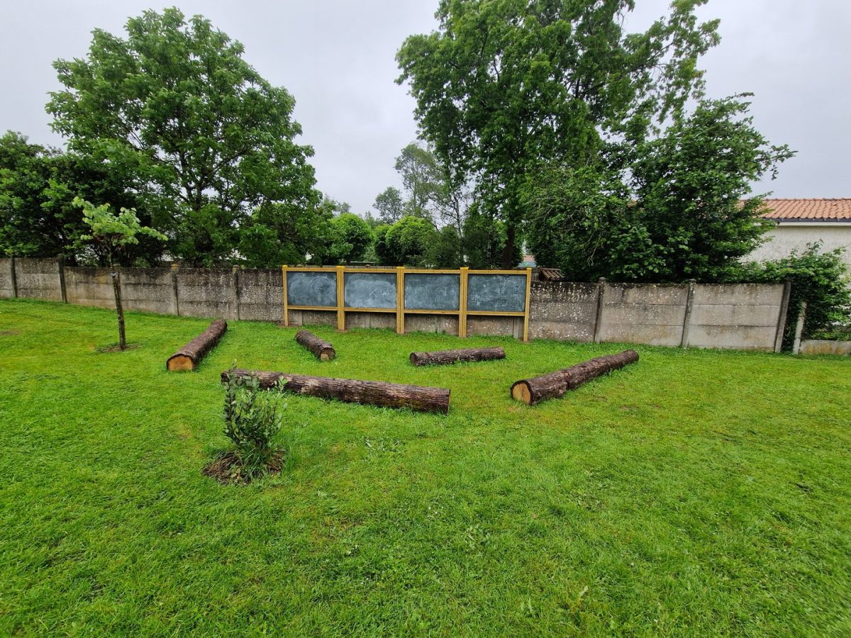 Espace pédagogique extérieur avec tableaux noirs sur mur béton, rondins de bois disposés sur pelouse verte