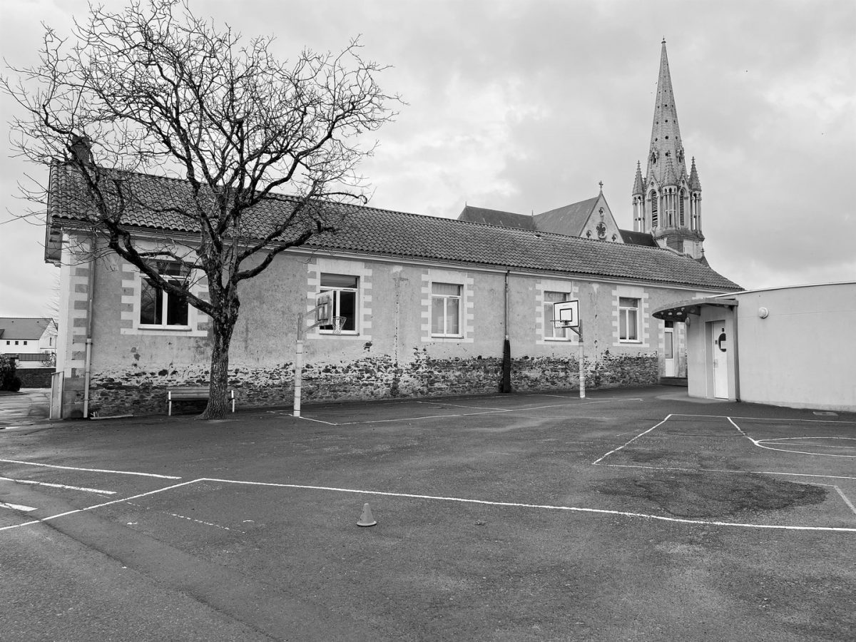 École en pierre avec cour de récréation, arbre dénudé au premier plan et clocher d'église gothique en arrière-plan