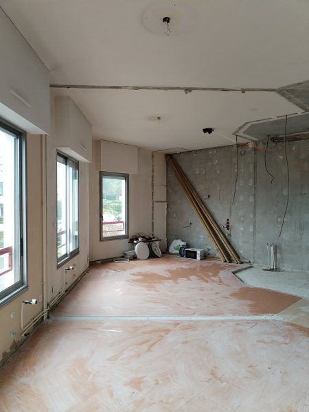 Espace intérieur en rénovation avec murs en béton brut, plancher en contreplaqué et grandes fenêtres