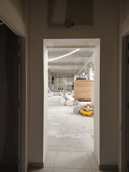 Vue depuis un couloir vers un espace commercial en chantier avec échafaudage, plafond béton apparent et luminaires suspendus