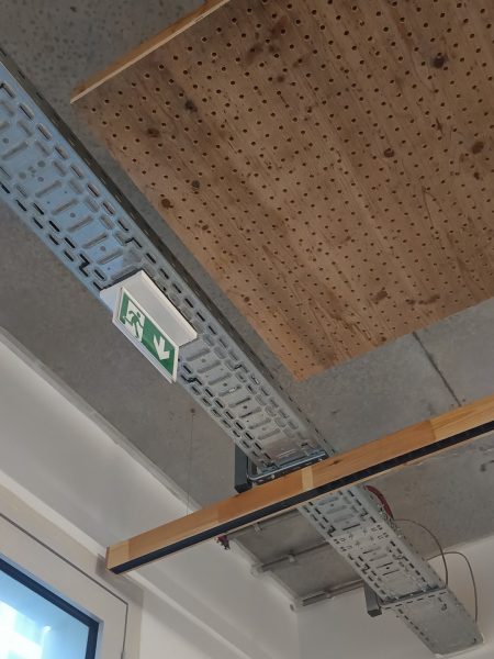 Plafond en béton avec panneau acoustique perforé en bois, câblage apparent et signalétique de sortie de secours