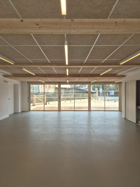 intérieur de la salle multiactivités avec vue sur le jardin par des grandes baies vitrées, les lumières sont allumées, la charpente apparente